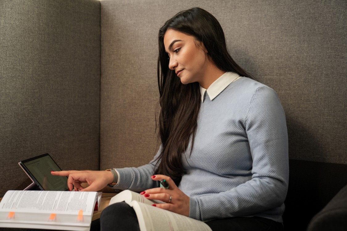 Junge Frau sitzt auf einem Sessel und liest am Tablet, während sie zwei Bücher auf dem Schoß aufgeschlagen hat. 